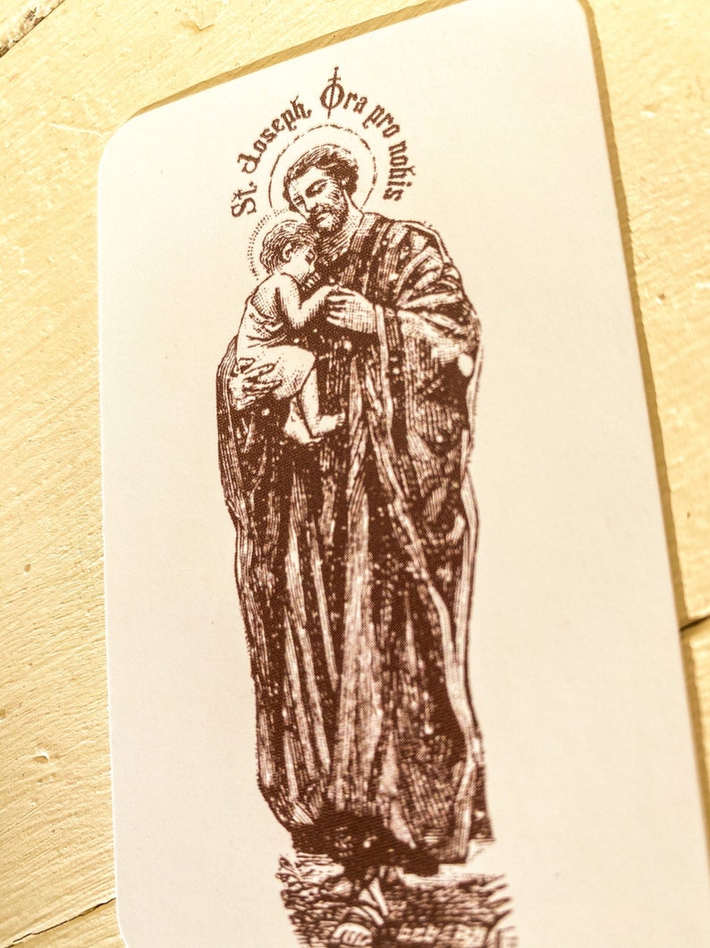 May include: A brown and white religious card with an illustration of Saint Joseph holding a baby Jesus. The card has the text "St. Joseph Ora pro nobis" in a circle around the image.
