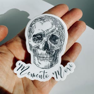 Puede incluir: Una pegatina troquelada que presenta una ilustración en tinta negra y blanca de un cráneo humano. Debajo del cráneo, el texto Memento Mori está impreso en una caligrafía cursiva negra. La pegatina tiene un borde blanco y acabado brillante.