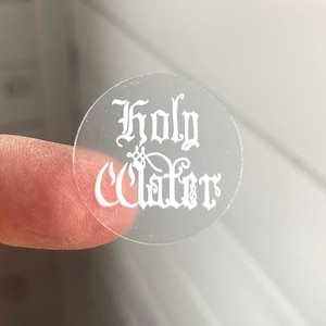 May include: A clear round sticker with the words "Holy Water" printed in white in a gothic font.