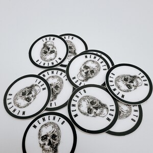Pode incluir: Uma cole&ccedil;&atilde;o de adesivos circulares com uma ilustra&ccedil;&atilde;o em preto e branco estilo tinta de um cr&acirc;nio humano no centro. O texto MEMENTO MORI est&aacute; impresso em uma fonte negrito sem serifa ao redor do per&iacute;metro de cada adesivo em fundo branco.