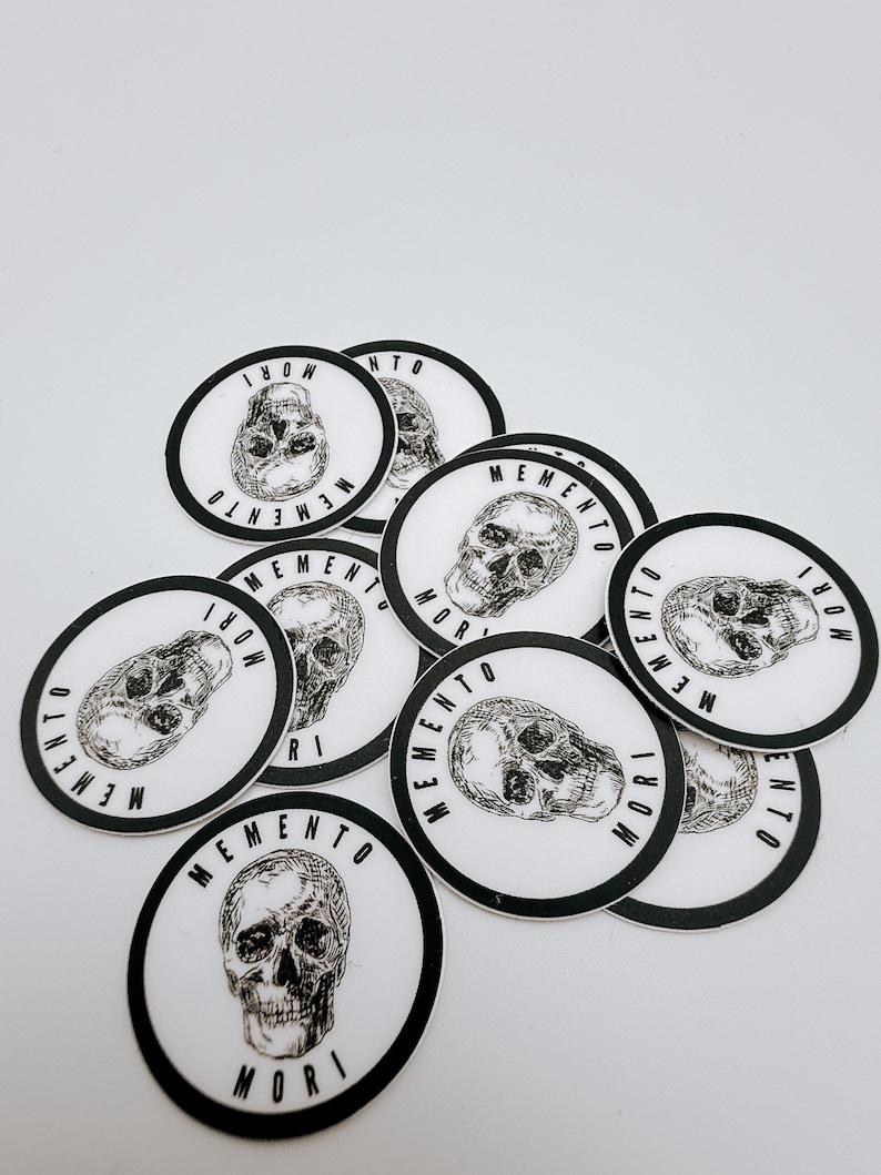 Pode incluir: Uma cole&ccedil;&atilde;o de adesivos circulares com uma ilustra&ccedil;&atilde;o em preto e branco estilo tinta de um cr&acirc;nio humano no centro. O texto MEMENTO MORI est&aacute; impresso em uma fonte negrito sem serifa ao redor do per&iacute;metro de cada adesivo em fundo branco.