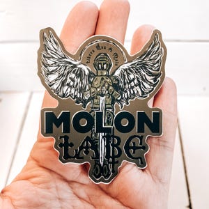 May include: A sticker featuring a winged angel holding a sword. The angel is wearing armour and has a halo. The sticker reads "Molon Labe" in black lettering.