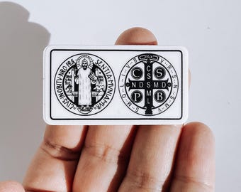 Pode incluir: Um adesivo retangular branco com uma impressão preta da medalha de São Bento. O lado esquerdo mostra uma figura segurando uma cruz e um livro, enquanto o lado direito exibe uma cruz com as letras C S P B, C S M L, N D S M D e PAX.