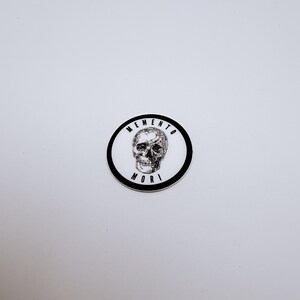 Pode incluir: Um autocolante circular com uma ilustra&ccedil;&atilde;o a preto e branco de um cr&acirc;nio humano centrado num fundo branco. O texto MEMENTO MORI est&aacute; impresso em letras mai&uacute;sculas pretas ao longo da margem interior da espessa borda circular preta.