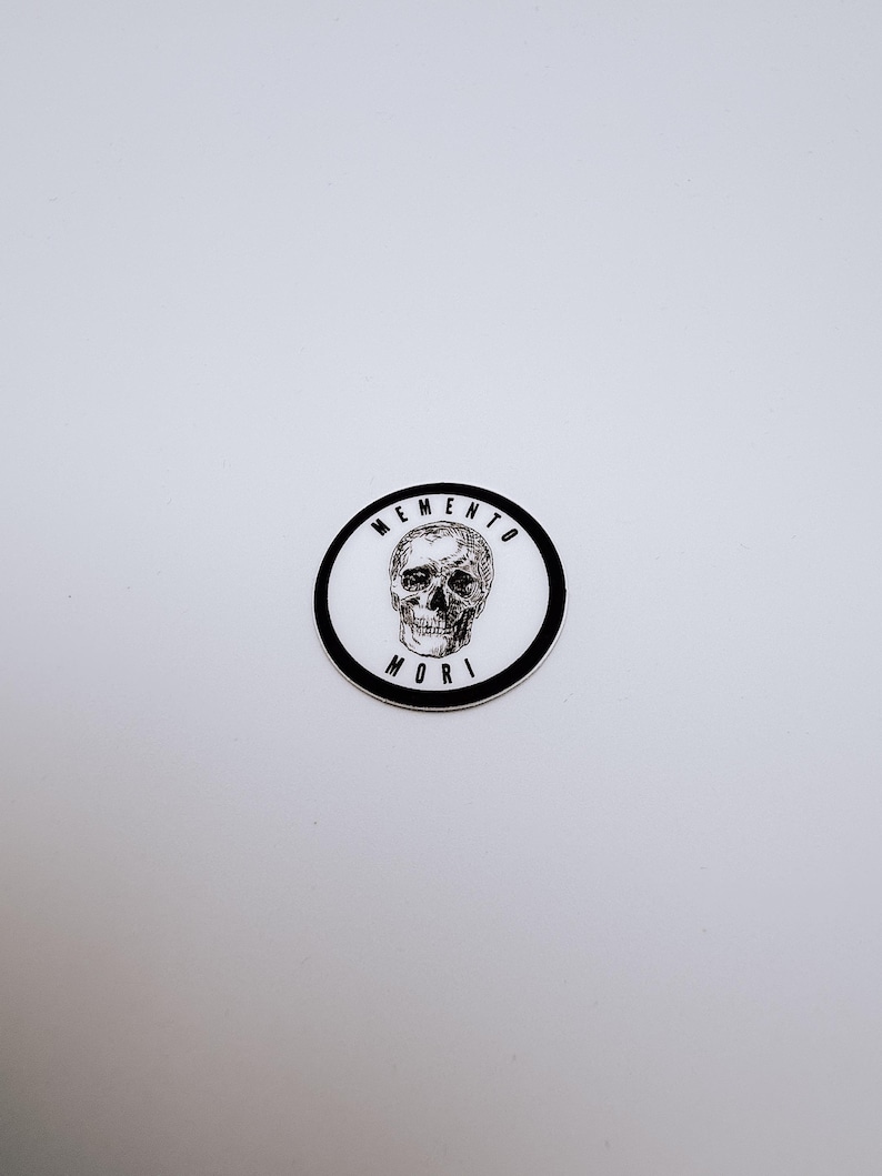Pode incluir: Um autocolante circular com uma ilustra&ccedil;&atilde;o a preto e branco de um cr&acirc;nio humano centrado num fundo branco. O texto MEMENTO MORI est&aacute; impresso em letras mai&uacute;sculas pretas ao longo da margem interior da espessa borda circular preta.