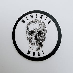Pode incluir: Um autocolante circular com uma ilustra&ccedil;&atilde;o a preto e branco de um cr&acirc;nio humano centrado num fundo branco. O texto MEMENTO est&aacute; impresso a preto num arco acima do cr&acirc;nio, e o texto MORI est&aacute; impresso a preto num arco abaixo.