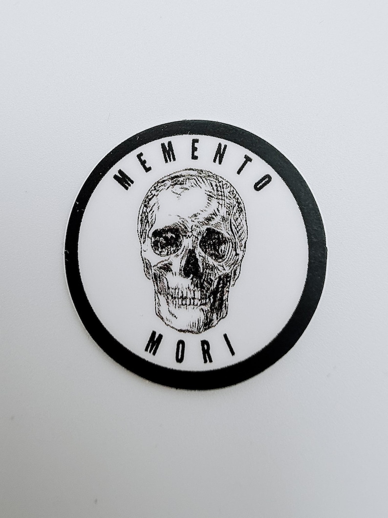 Pode incluir: Um autocolante circular com uma ilustra&ccedil;&atilde;o a preto e branco de um cr&acirc;nio humano centrado num fundo branco. O texto MEMENTO est&aacute; impresso a preto num arco acima do cr&acirc;nio, e o texto MORI est&aacute; impresso a preto num arco abaixo.