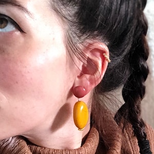 Peut inclure: Une femme avec de longs cheveux bruns portant un pull à col roulé marron et une paire de boucles d'oreilles de forme ovale jaune et marron.