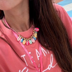 Peut inclure: Boucles d'oreilles roses en forme de fleur et collier de coquillages colorés avec des accents dorés. Le collier présente une variété de coquillages aux couleurs pastel. Un t-shirt rose avec le texte "TEAM Bride" est également visible.