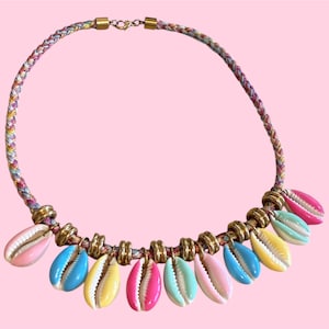 Könnte beinhalten: Eine farbenfrohe Halskette mit einem geflochtenen Band in Rosa-, Gelb- und Blautönen. Die Halskette ist mit mehreren Muschelanhängern in verschiedenen Pastellfarben verziert, die durch goldfarbene Metallringe verbunden sind.