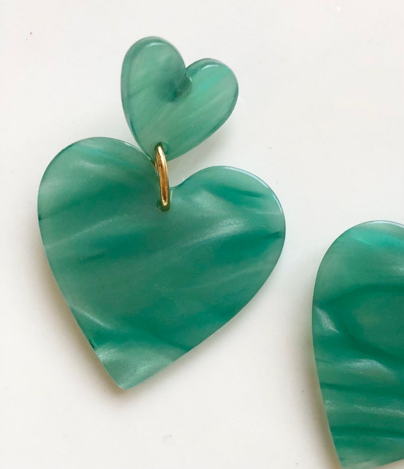 May include: A pair of green heart-shaped earrings with a gold accent. The earrings are made of a marbled material.