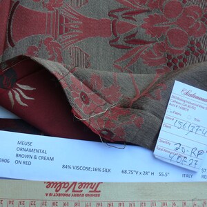 May include: A close-up of a red and brown fabric with a floral pattern. The fabric is labeled "Meuse Ornamental Brown & Cream on Red" and "84% Viscose; 16% Silk". The fabric is 68.75 inches wide by 28 inches high and has a 55.5 inch repeat. The fabric is made in Italy.