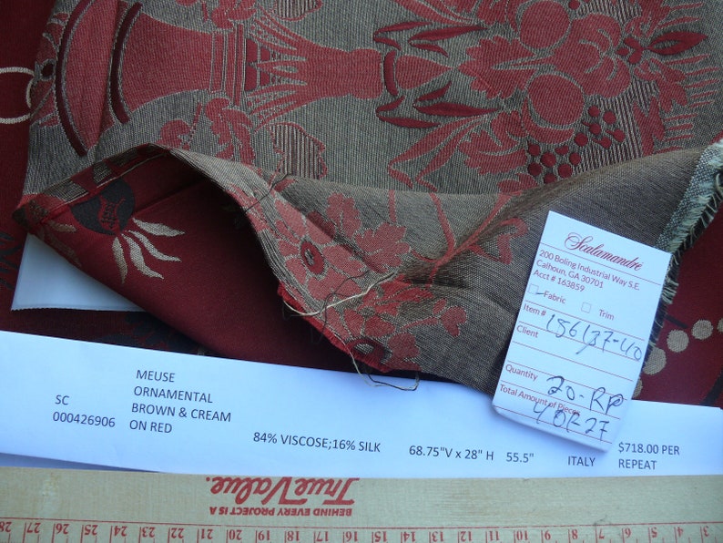 May include: A close-up of a red and brown fabric with a floral pattern. The fabric is labeled "Meuse Ornamental Brown & Cream on Red" and "84% Viscose; 16% Silk". The fabric is 68.75 inches wide by 28 inches high and has a 55.5 inch repeat. The fabric is made in Italy.
