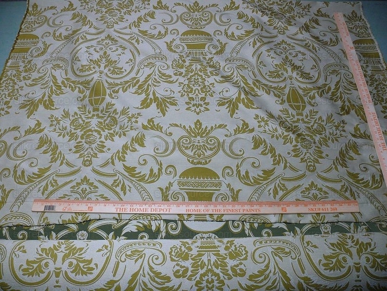 Pu&ograve; includere: Un primo piano di un tessuto bianco con un motivo damascato dorato. Il tessuto ha una lucentezza sottile e una texture morbida. Il motivo presenta intricati motivi floreali e disegni ripetitivi. Il tessuto &egrave; piegato a met&agrave; e un righello &egrave; posto sopra.