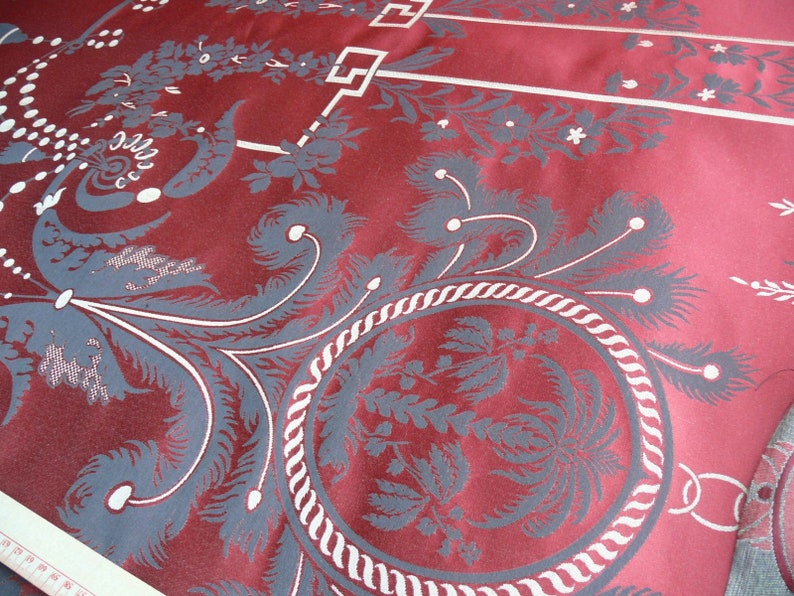 May include: Close-up of a red fabric with a white and gray floral pattern. The fabric has a textured, woven look and features a repeating design of stylized flowers and leaves.