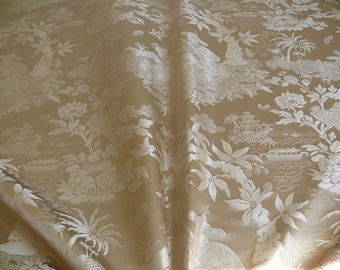 Pode incluir: Um tecido dourado com um padrão floral branco. O padrão apresenta flores, folhas e pássaros estilizados. O tecido tem um brilho sutil.
