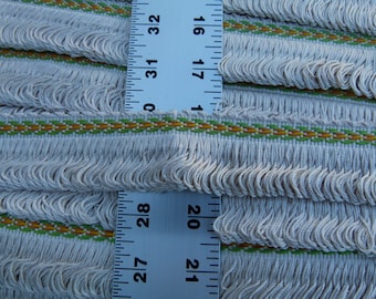 May include: Close-up of white fringe trim with a green and gold woven stripe. The trim is measured with a ruler showing 27 to 32 inches.
