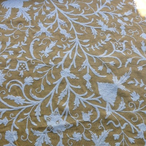 May include: A beige fabric with a white embroidered floral pattern. The design features intricate vines, leaves, and flowers.