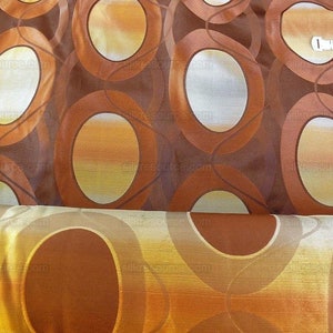 以下が含まれることがあります： 茶色と金の楕円形の模様が繰り返される生地のクローズアップ。生地は半分に折り畳まれており、両側に模様が見えます。