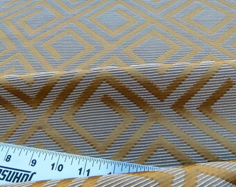 May include: Close-up of a fabric with a geometric pattern. The design features repeating diamond shapes in shades of gold and gray. The fabric has a textured appearance, with a ruler showing inches. This textile is suitable for various craft projects.