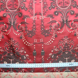 May include: Red fabric with a black and white damask pattern. The pattern features a repeating design of stylized trees, flowers, and geometric shapes. A ruler is placed on the fabric to show the scale.