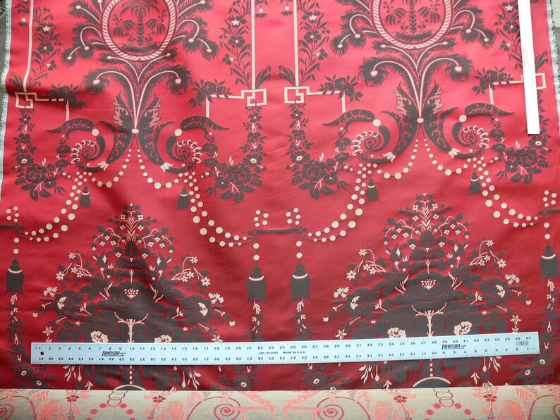 May include: Red fabric with a black and white damask pattern. The pattern features a repeating design of stylized trees, flowers, and geometric shapes. A ruler is placed on the fabric to show the scale.