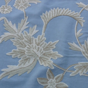 May include: A light blue fabric with a white embroidered floral design. The design features delicate leaves and flowers.