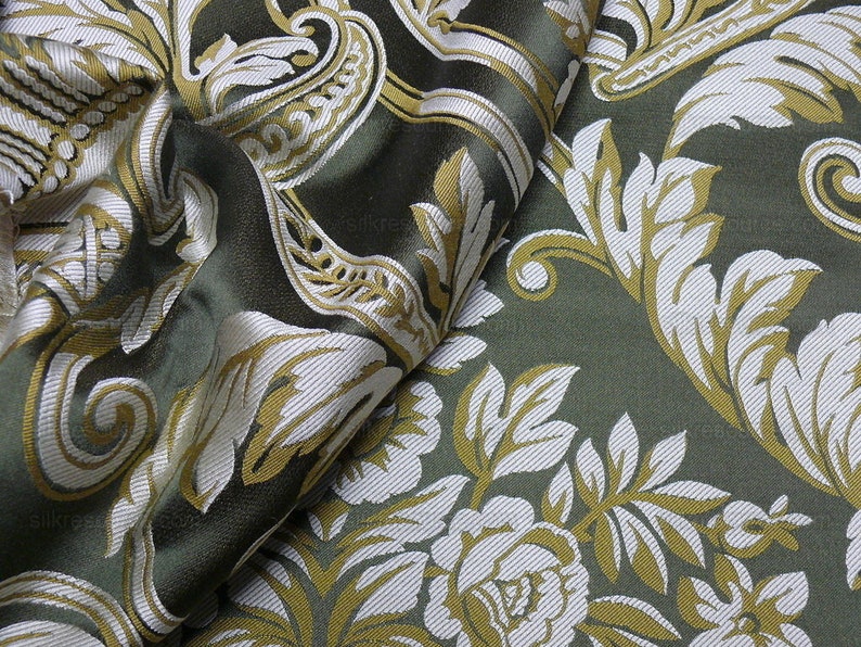 Pu&ograve; includere: Primo piano di un tessuto verde con un motivo floreale bianco e oro. Il tessuto &egrave; testurizzato e presenta un design simile al damasco.