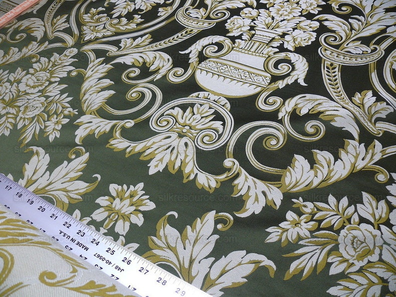 Pu&ograve; includere: Tessuto verde con un motivo floreale bianco e oro. Il motivo presenta un disegno ripetitivo di fiori stilizzati, foglie e un vaso con fiori.