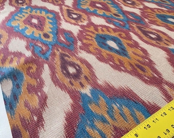 May include: Close-up of a fabric with a colourful ikat pattern. The pattern features shades of red, blue, yellow, and brown. The fabric has a woven texture.