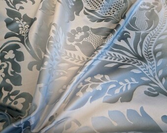 Può includere: Un tessuto blu chiaro con un motivo damascato bianco. Il tessuto è piegato e drappeggiato, mostrando i dettagli intricati del design.