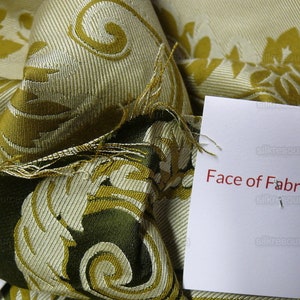Pu&ograve; includere: Primo piano di un campione di tessuto con un motivo damascato oro e verde. Il tessuto &egrave; piegato e un'etichetta bianca con il testo "Face of Fabric" &egrave; allegata.