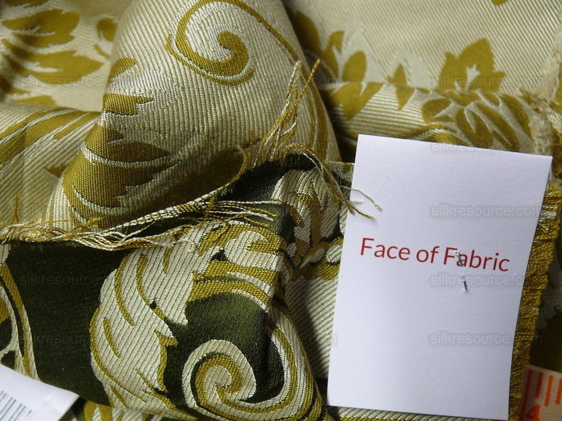 Pu&ograve; includere: Primo piano di un campione di tessuto con un motivo damascato oro e verde. Il tessuto &egrave; piegato e un'etichetta bianca con il testo "Face of Fabric" &egrave; allegata.