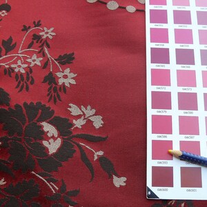 May include: A close-up of a red fabric with a floral pattern in black and white. A color chart with a pencil is on the right side of the image. The color chart has a variety of pink and red shades.