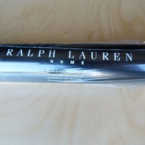 Puede incluir: Un rollo de papel tapiz enrollado con una etiqueta negra que dice "RALPH LAUREN HOME" y "RALPH LAUREN HOME, AN EXCLUSIVE COLLECTION OF FURNISHINGS FOR THE HOME, A TRIUMPH OF AMERICAN DESIGN."