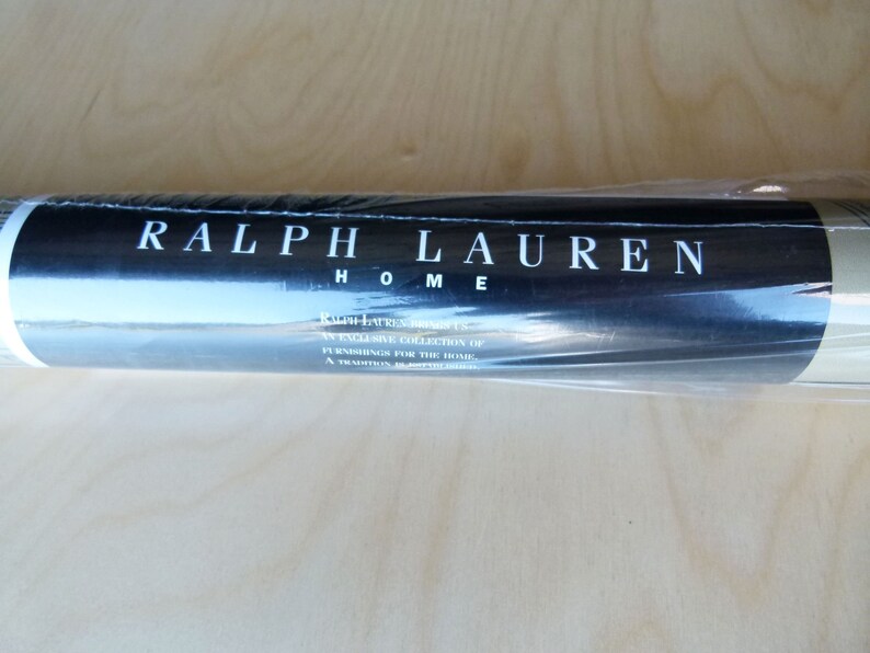 Puede incluir: Un rollo de papel tapiz enrollado con una etiqueta negra que dice "RALPH LAUREN HOME" y "RALPH LAUREN HOME, AN EXCLUSIVE COLLECTION OF FURNISHINGS FOR THE HOME, A TRIUMPH OF AMERICAN DESIGN."