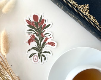 May include: A sticker featuring a stylized illustration of a plant with red and green leaves. The sticker is on a white surface, next to a cup of tea, a decorative book, and dried plants.