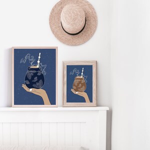 Peut inclure: Deux impressions encadrées d'une boisson dans une tasse en forme de gourde avec une paille, tenue dans une main, sur fond bleu foncé. Les impressions sont exposées sur une étagère blanche, avec un chapeau de paille suspendu au-dessus. L'œuvre est de style minimaliste.