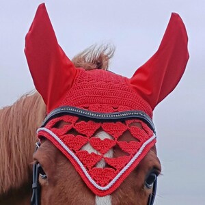 May include: A chestnut horse wearing a red crochet fly bonnet with white accents and a black bridle. The horse is in a field with green grass under an overcast sky. The bonnet covers the horse's ears and forehead.
