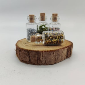 May include: Four small glass bottles with cork stoppers, each filled with different decorative elements. One bottle is lying on its side, filled with gold glitter. The other bottles contain silver, green, and multicolored materials. The bottles are displayed on a wooden slice.