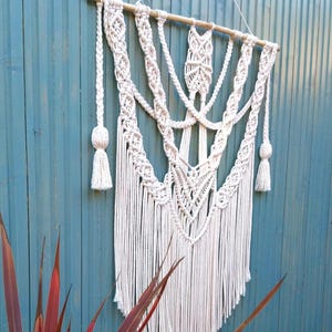 May include: Cream-colored macrame wall hanging with intricate woven patterns and fringe. The textile art piece is suspended from a wooden dowel and hangs against a teal blue wooden fence. Decorative tassels are on each side.
