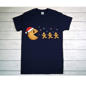 May include: Navy blue t-shirt featuring a humorous Christmas design. A Pac-Man-like cookie wearing a Santa hat is chasing gingerbread men. Snowflakes and candy canes complete the festive scene. A fun, holiday-themed graphic tee.