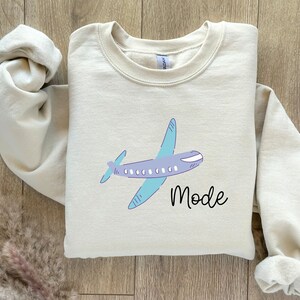 Op de afbeelding: Een beige sweatshirt met een cartoon vliegtuig en het woord "Mode" erop gedrukt.