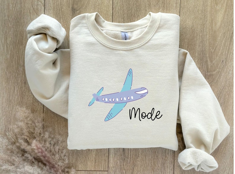 Op de afbeelding: Een beige sweatshirt met een cartoon vliegtuig en het woord "Mode" erop gedrukt.