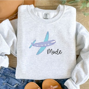 Op de afbeelding: Een grijs sweatshirt met een cartoon vliegtuig en het woord "Mode" erop gedrukt.