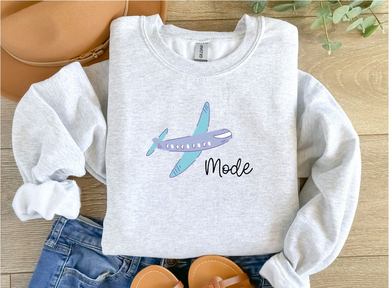 Op de afbeelding: Een grijs sweatshirt met een cartoon vliegtuig en het woord "Mode" erop gedrukt.
