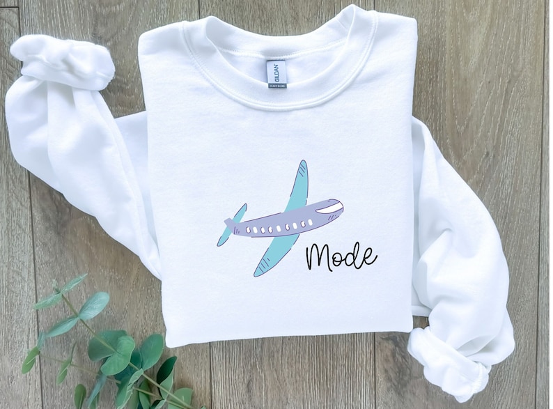 Op de afbeelding: Witte trui met een paarse en blauwe vliegtuiggrafiek en de tekst "Mode" gedrukt op de voorkant.