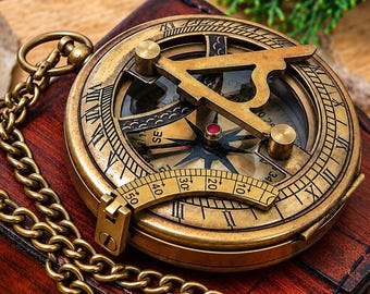 May include: A brass sundial compass with a hinged gnomon and a curved scale, featuring Roman numerals and cardinal directions on its face. The circular instrument is attached to a gold-toned metal chain and rests upon a dark red wooden box.