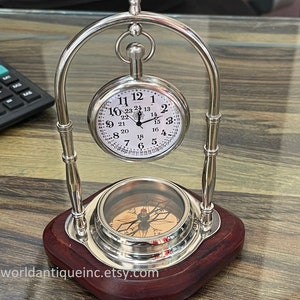 May include: A silver pocket watch with a compass on a wooden stand. The watch face has black Roman numeral markings and a silver hand. The compass has a red needle and a decorative design.