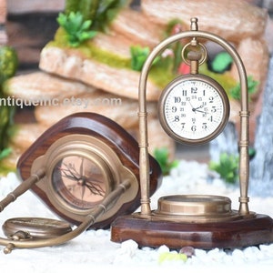 May include: A brass compass and pocket watch on a wooden stand. The compass has a white face with black markings and a red needle. The pocket watch has a white face with black numbers and hands. The stand is made of dark wood with a brass base.